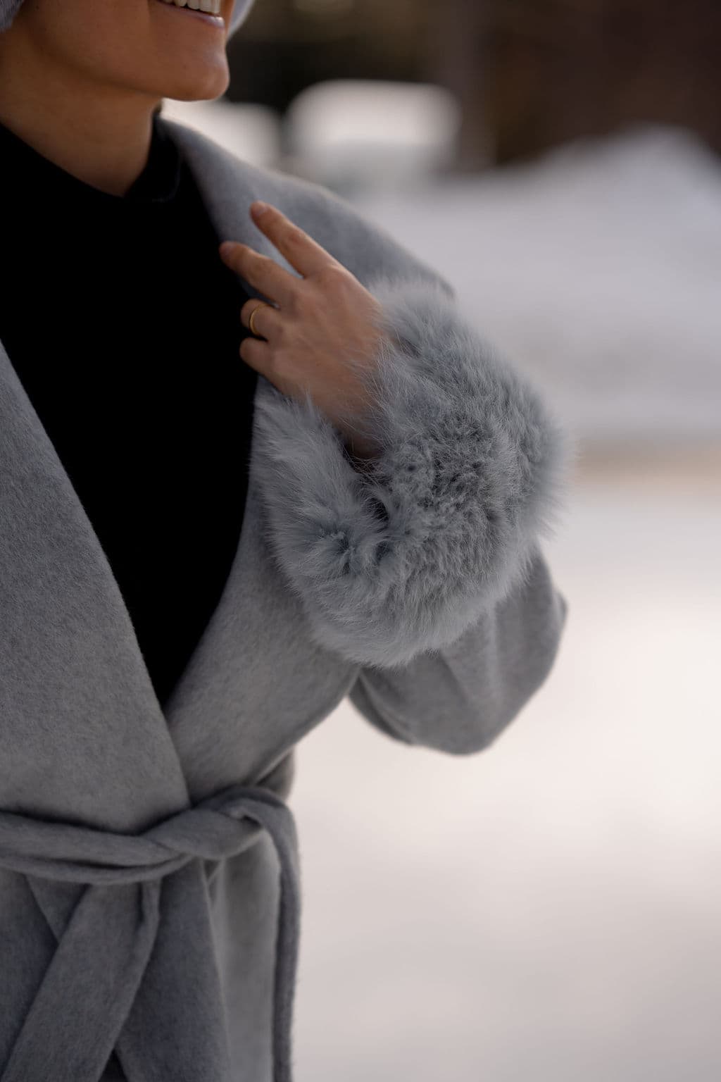 A belted grey wool coat, its shawl collar held at the chest, with a pale grey fox cuff.